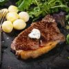 Grilled steak with butter, potatoes and green salad over black metal board